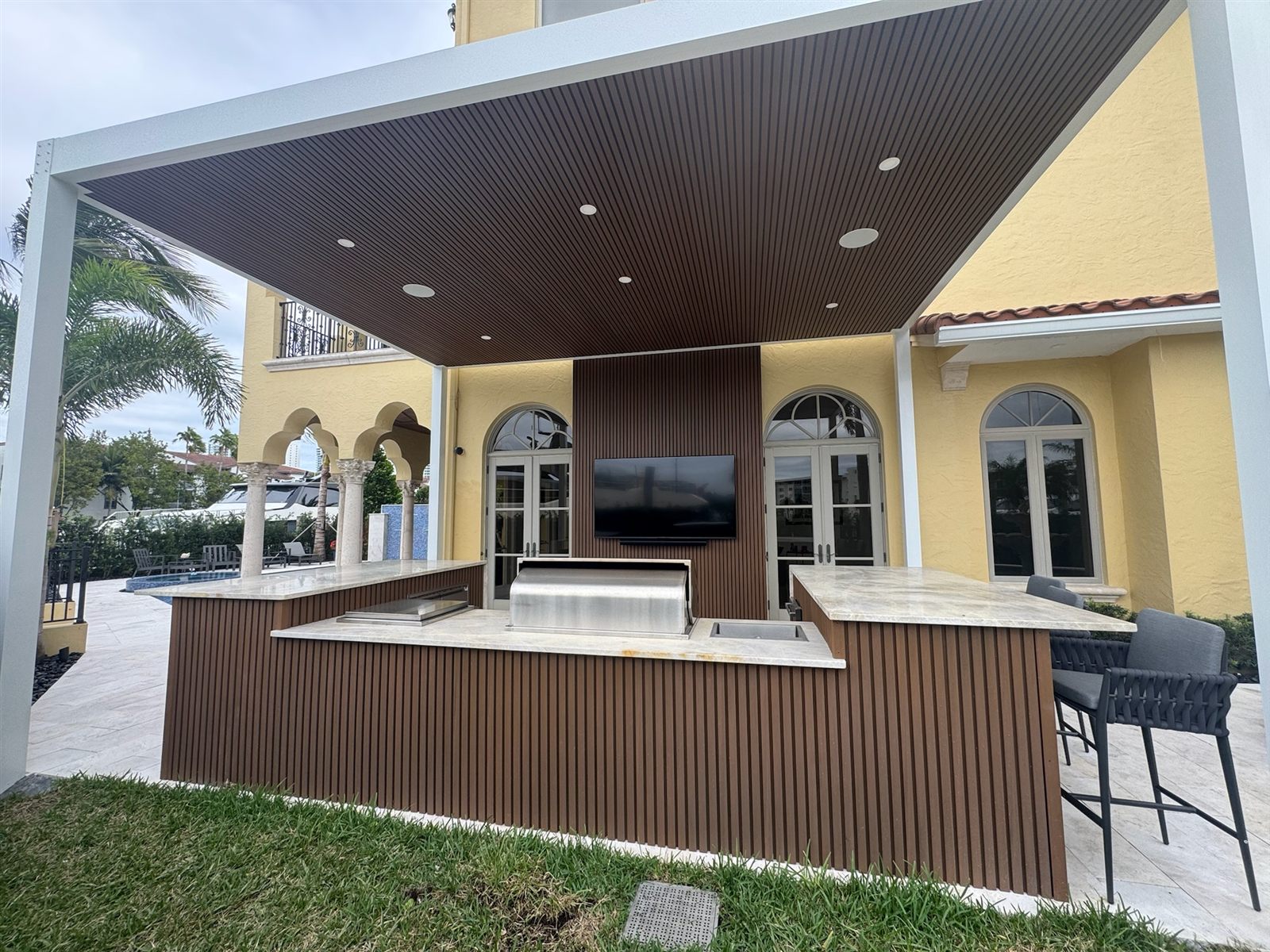 Underside of Open Sky pergola showing the fluted-wood ceiling with recessed lighting over an outdoor kitchen